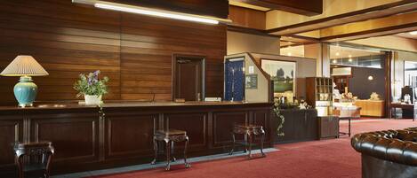A reception area with a wooden reception desk, two metal stools, a red carpet, and a leather couch.