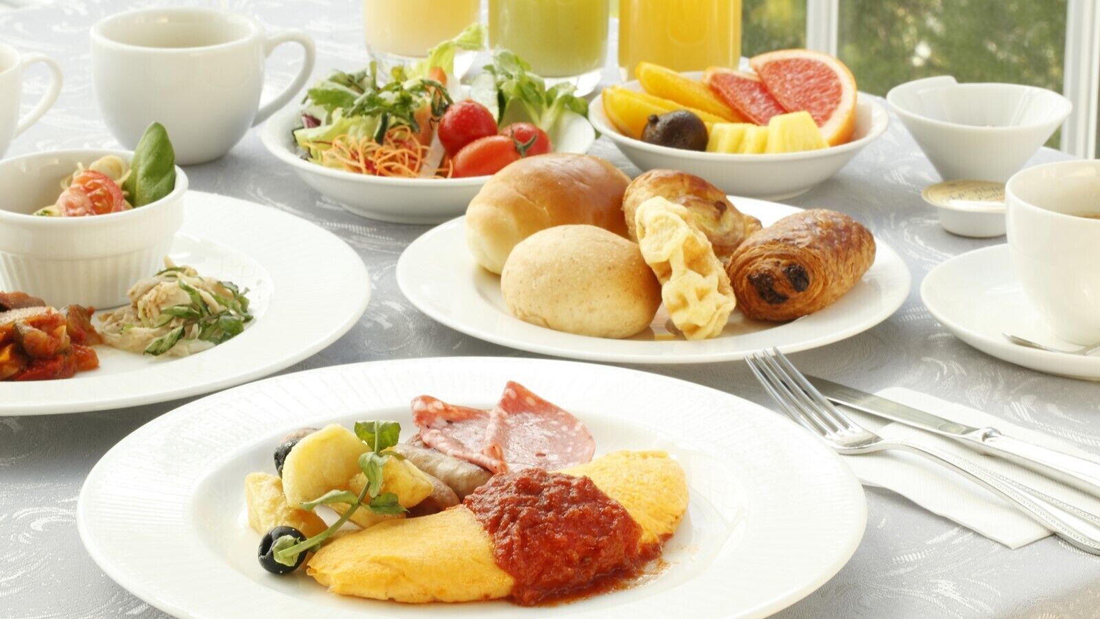 Une table dressée pour le petit-déjeuner, avec des assiettes remplies de différents aliments, notamment des œufs, du jambon et des viennoiseries, ainsi qu’une variété de fruits et de légumes.