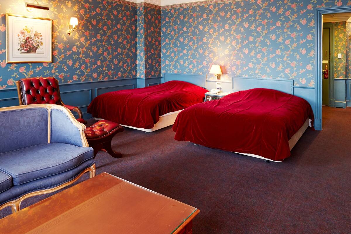 A room with two beds, a chair, a table, and a wall-mounted lamp.