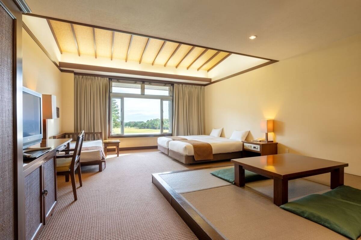 A hotel room with a bed, a desk with a computer, a chair, a table, and a window with curtains.