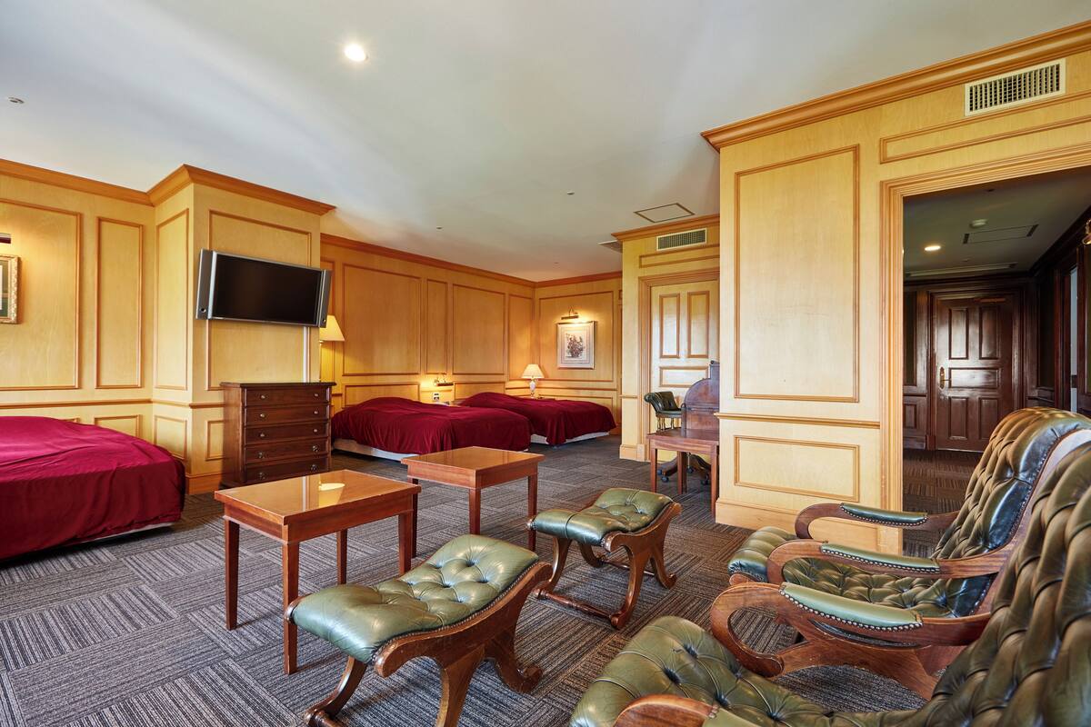 A hotel room with a large bed, a desk, two armchairs, a TV, and a door leading to another room.