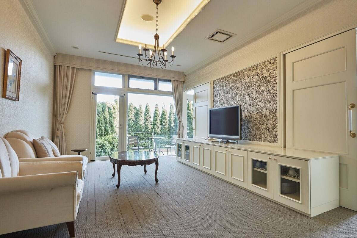 A spacious room with a sofa, a glass-top dining table, a TV on a cabinet, and a view of trees through large windows.