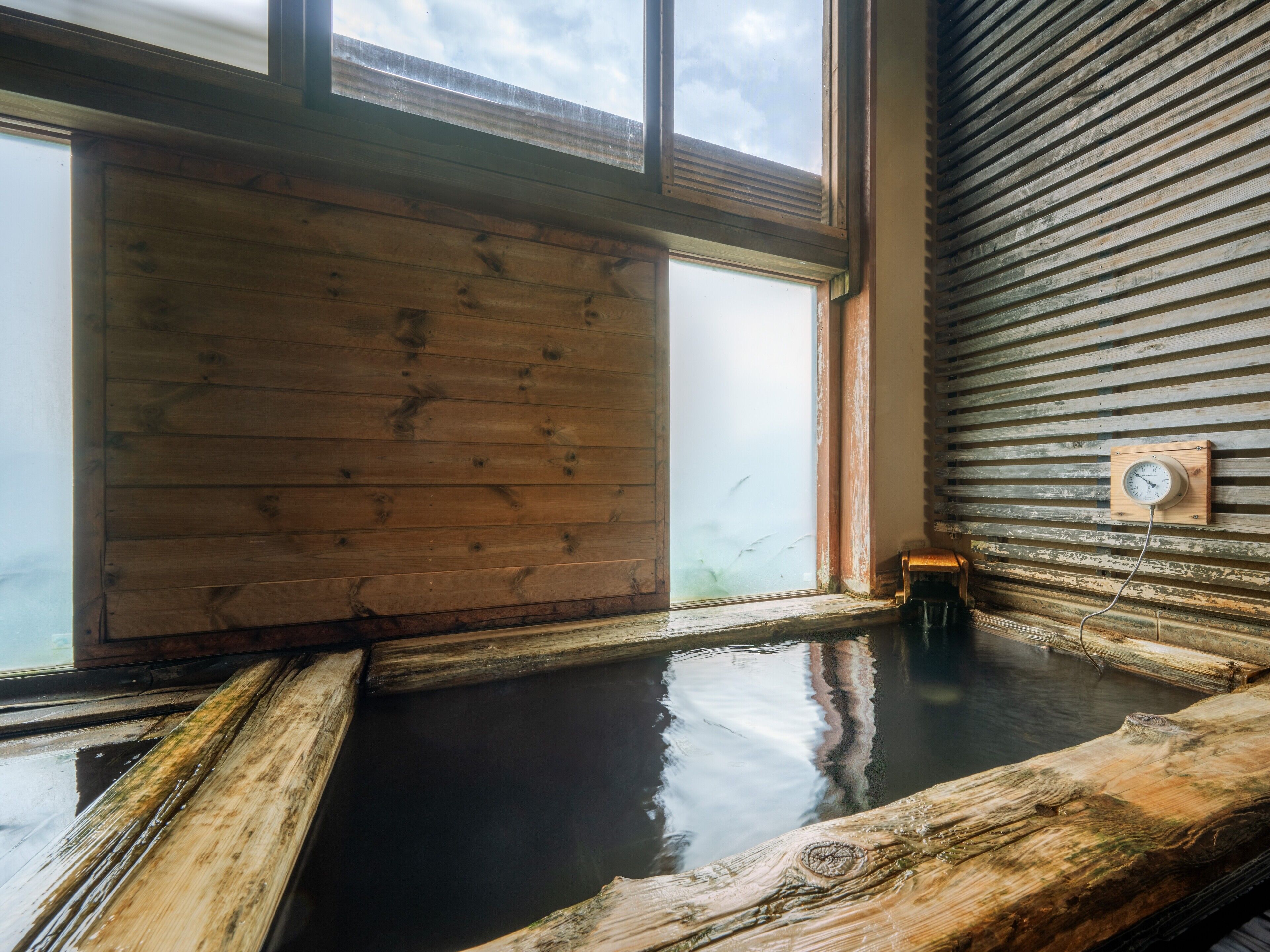 A wooden interior with a large window, a wooden bench, and a wall-mounted thermometer.