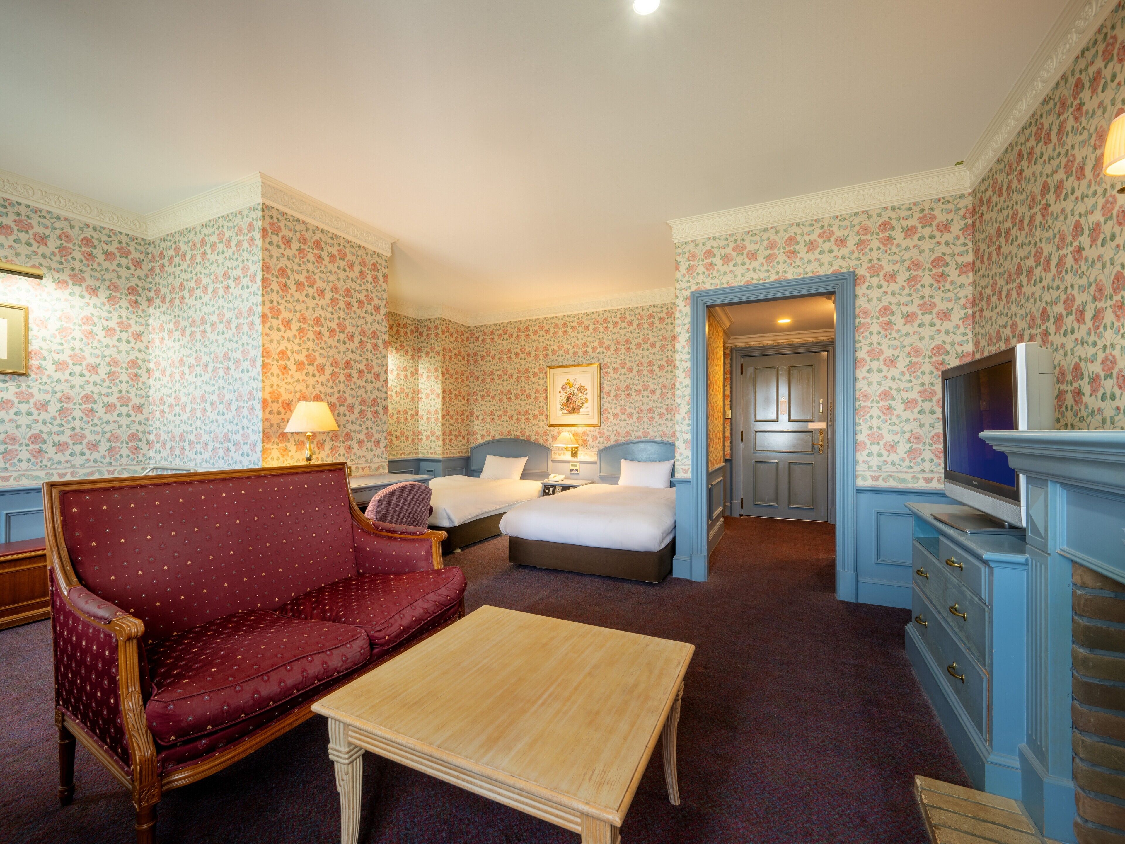 Une chambre d’hôtel avec un papier peint à motifs floraux, un canapé rouge recouvert de tissu, deux lits, une table basse en bois, une télévision et une commode.