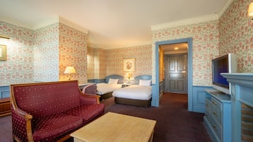 A hotel room with floral wallpaper, a red upholstered sofa, two beds, a wooden coffee table, a television, and a dresser.