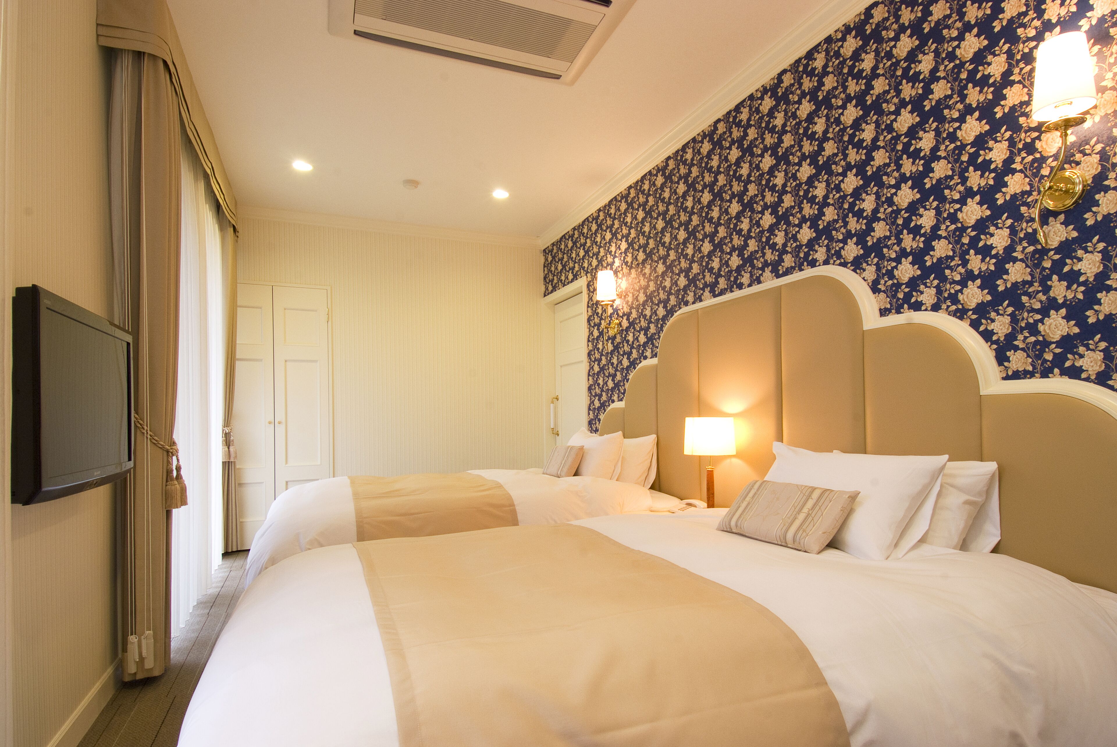 A hotel room with two beds, a television, a wall-mounted lamp, and a floral wallpaper.