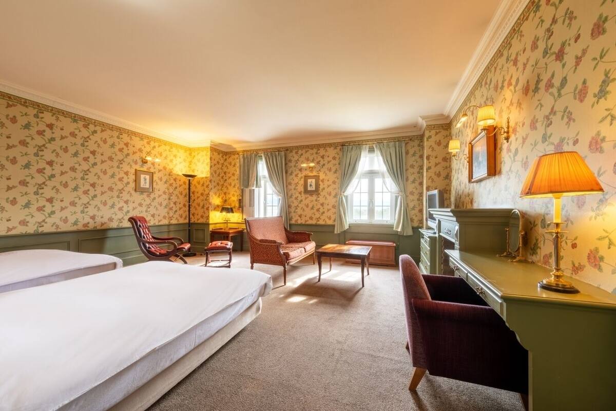 A hotel room with two beds, a desk, a chair, a TV, and a lamp.