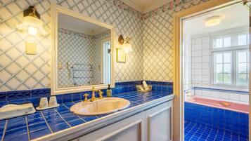 Un bagno con uno specchio grande, un lavandino e una vasca da bagno. Le pareti sono rivestite di piastrelle a motivo a diamante, e una finestra lascia entrare luce naturale.