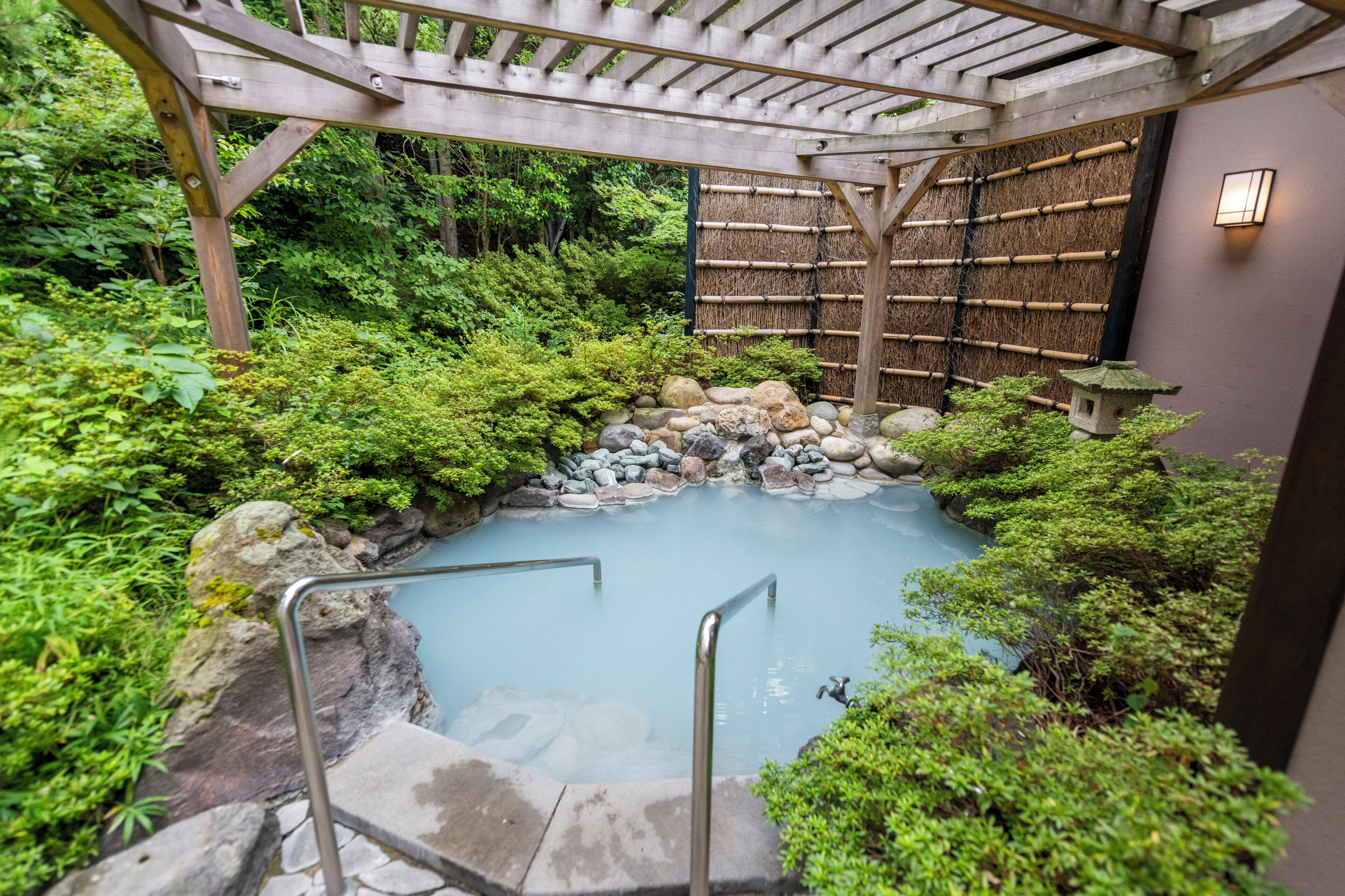 Une source thermale entourée d’une végétation luxuriante, une pergola en bois et un mur en bambou.