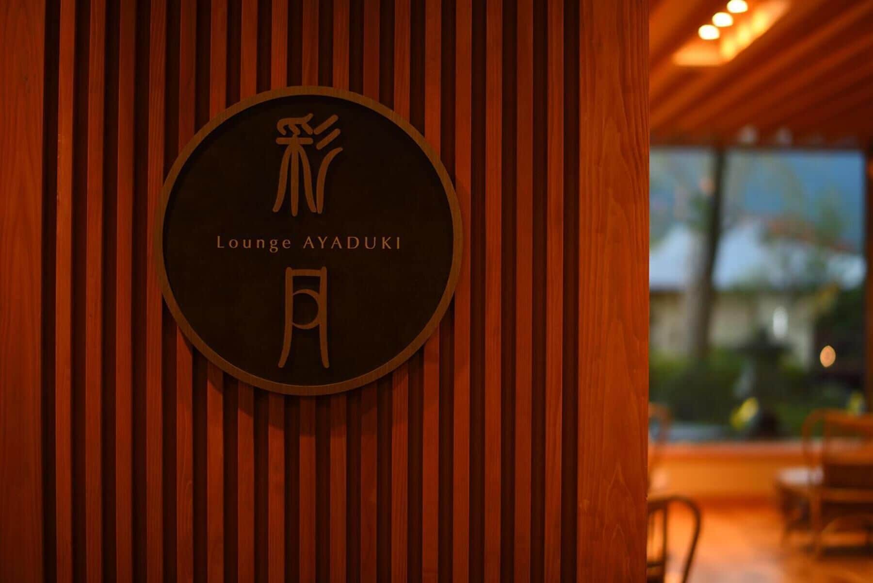 A circular sign with Japanese characters and the text "Lounge AYADUKI" on a wooden wall.