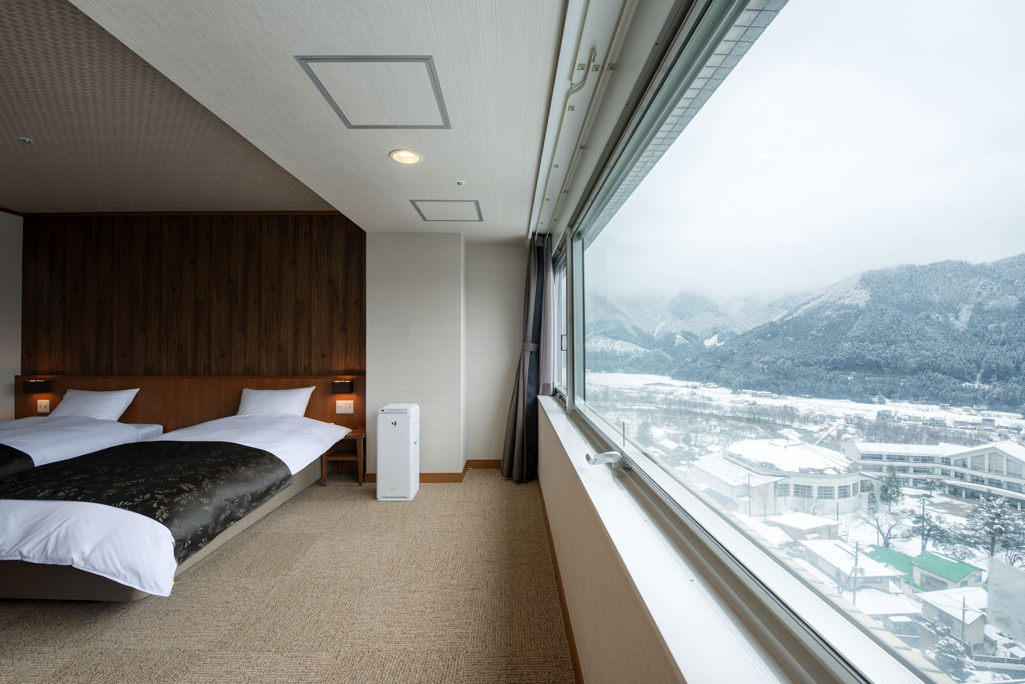 Quarto de hotel com duas camas, cabeceira de madeira, aquecedor portátil e vista para uma paisagem nevada através de uma janela grande.