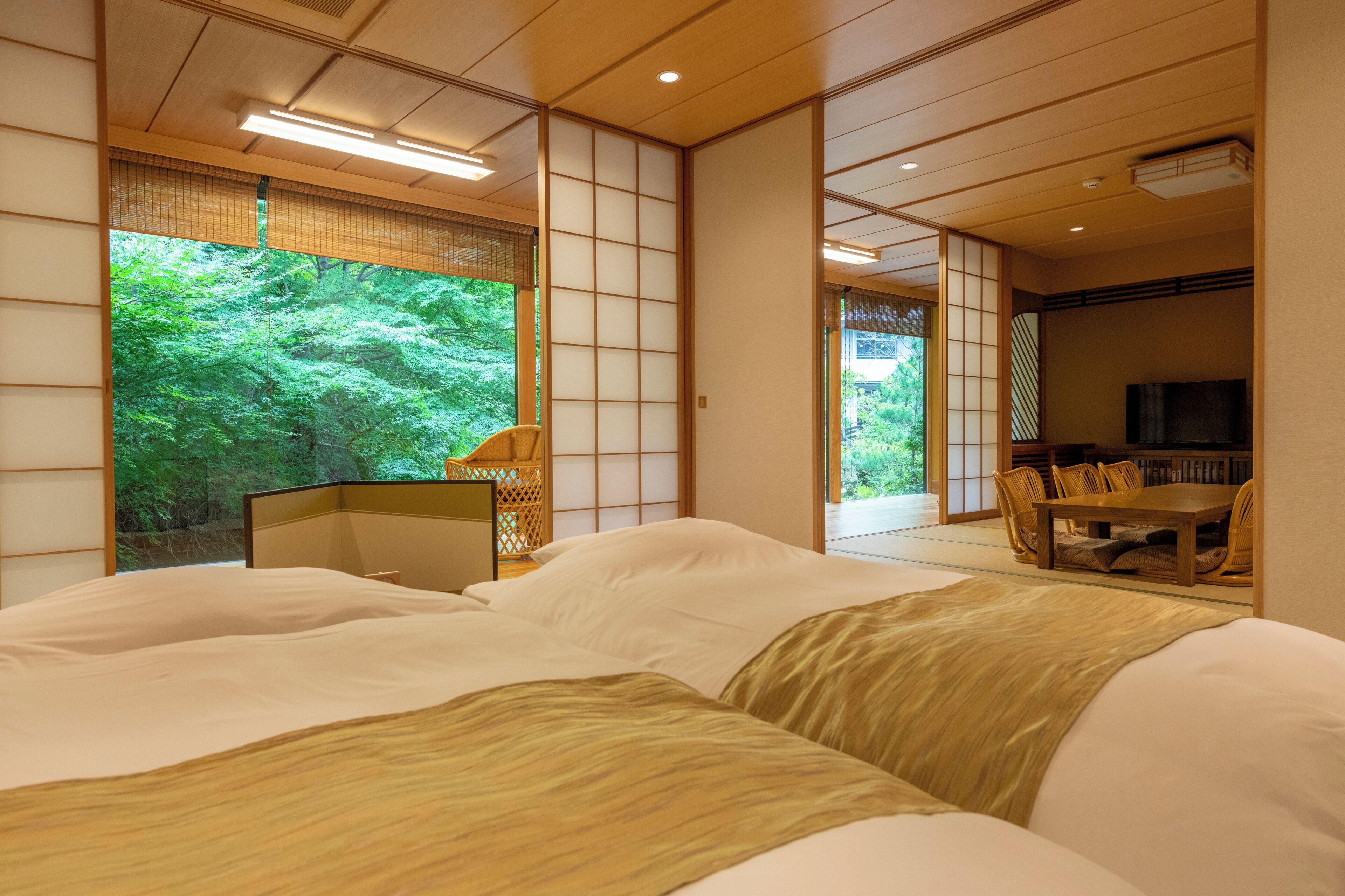 Un dormitorio de estilo japonés tradicional, con suelo de tatami, una mesa baja con sillas y un ventanal con vistas a la vegetación.