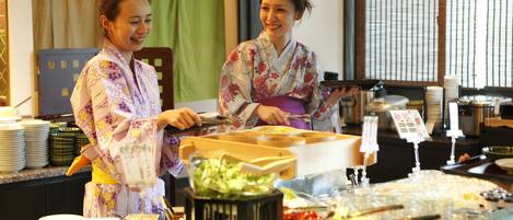Two individuals in traditional attire serving food at a buffet with various dishes and condiments.