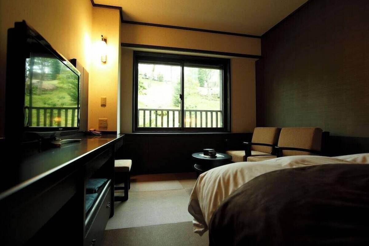 A hotel room with a bed, a desk with a computer, a chair, a small table, and a window with a view of greenery.