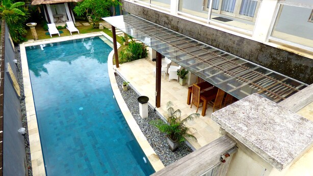 A swimming pool with a glass balustrade, surrounded by a garden and a thatched-roof structure. - Villa Mels (Jimbaran)