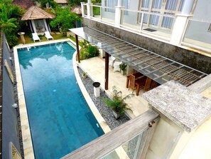 A swimming pool with a glass balustrade, surrounded by a garden and a thatched-roof structure. - Villa Mels (Jimbaran)