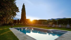 Una piscina rectangular con borde de piedra, rodeada por un césped cuidado y una fila de sillas.