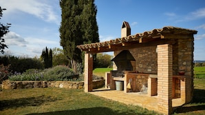 Una cocina al aire libre de piedra con chimenea, rodeada por un jardín con diversas plantas y un árbol alto.