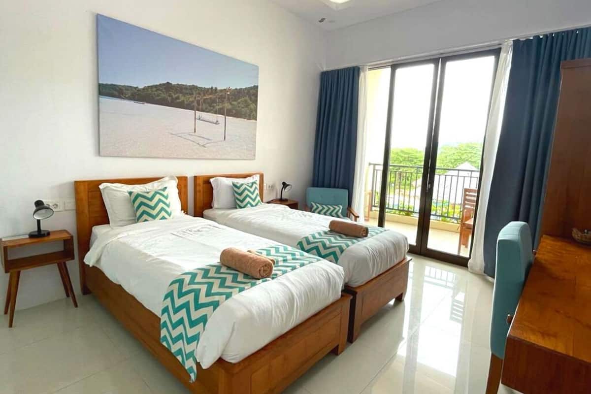 A hotel room with two beds, a desk, a chair, a balcony with a view, and a painting on the wall.