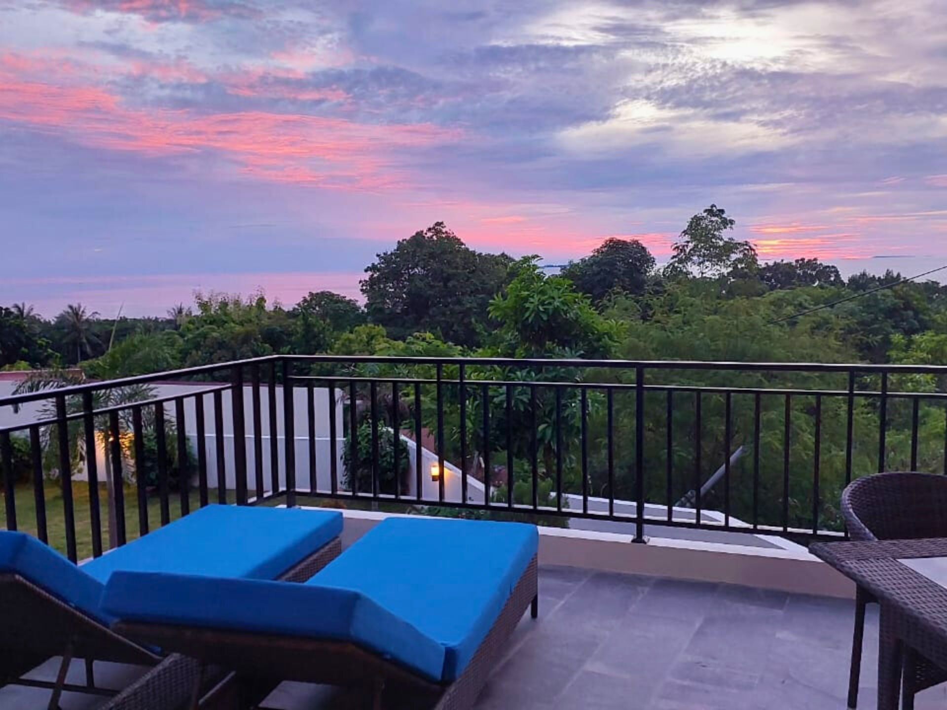 Un balcón con sillones de descanso azules y una mesa de mimbre, con vistas a un paisaje exuberante con árboles y un cielo al atardecer.