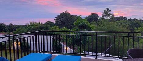 Un balcón con sillones de descanso azules y una mesa de mimbre, con vistas a un paisaje exuberante con árboles y un cielo al atardecer.