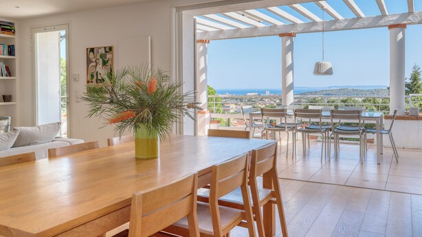 Een ruime eetruimte met een houten tafel en stoelen, een kamerplant en uitzicht op de stad en zee door grote ramen.