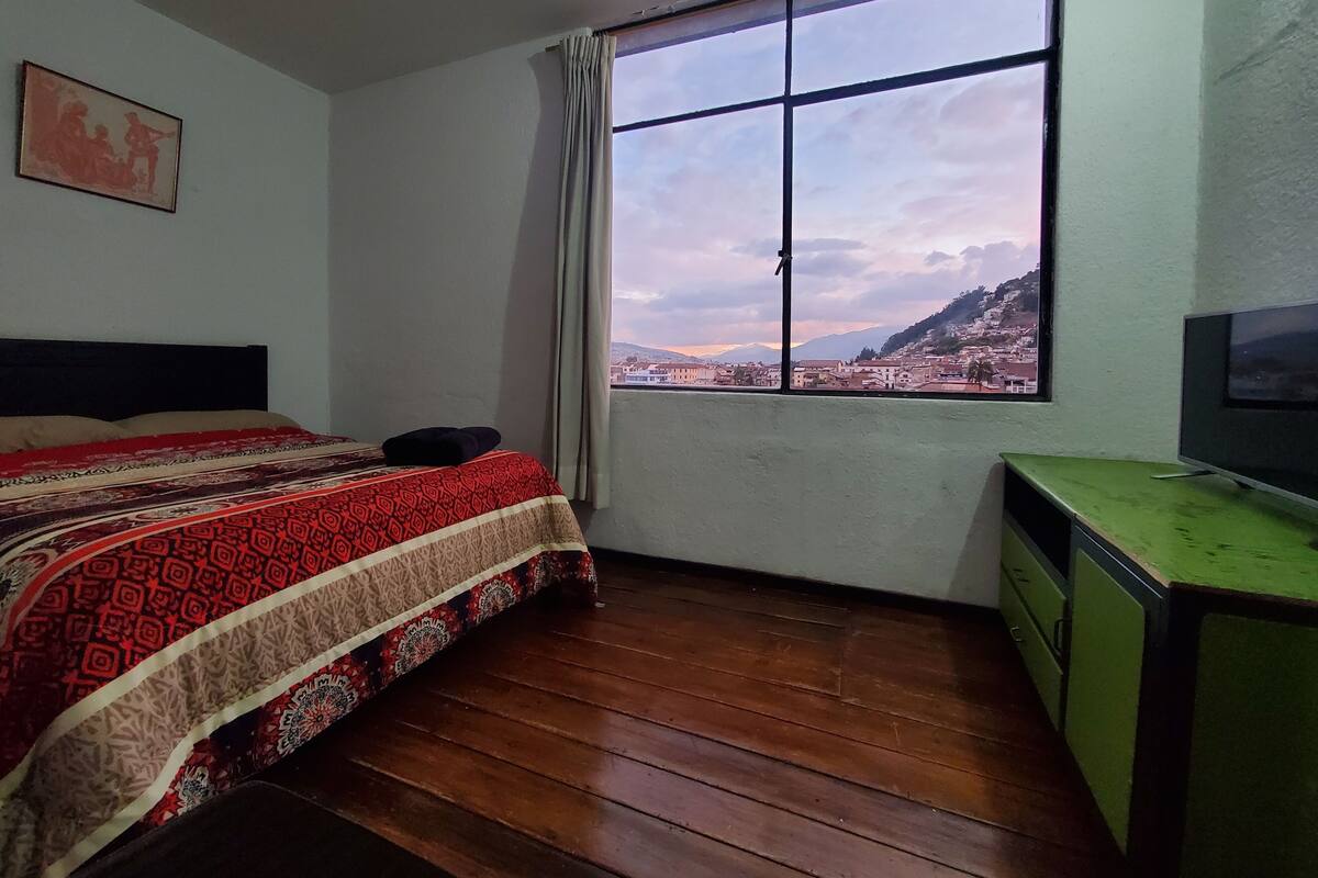 A bedroom with a bed, a window with a city view, a green desk, and a television.