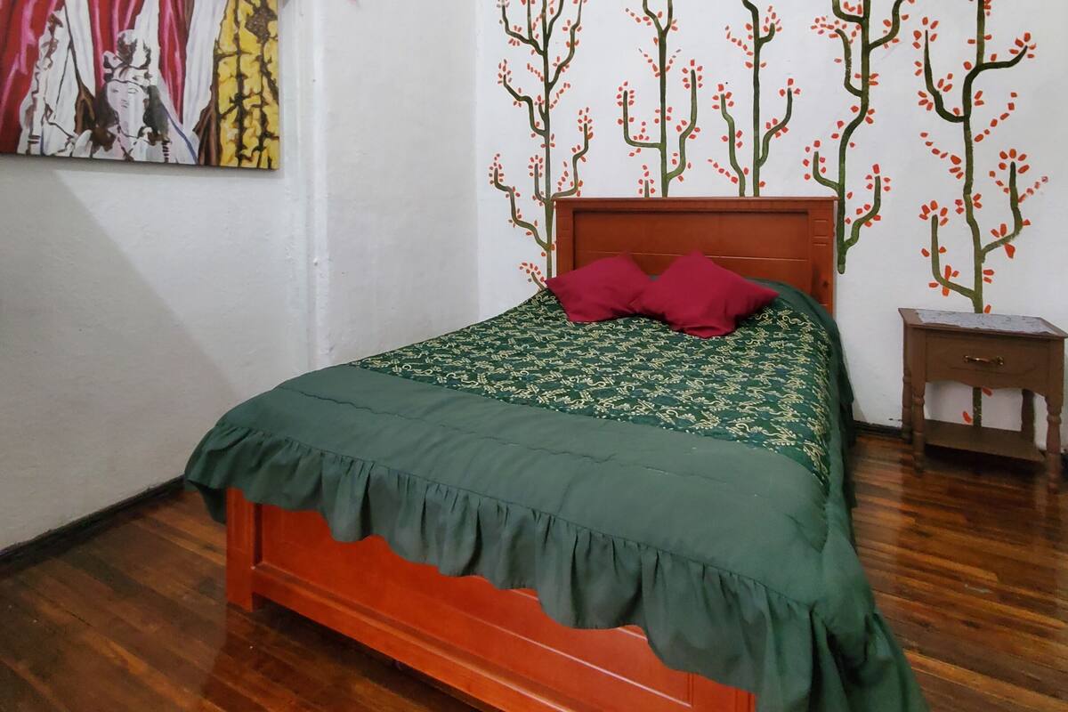 A bedroom with a wooden bed, a green bedspread, a red pillow, a small wooden nightstand, and a wall mural of trees.