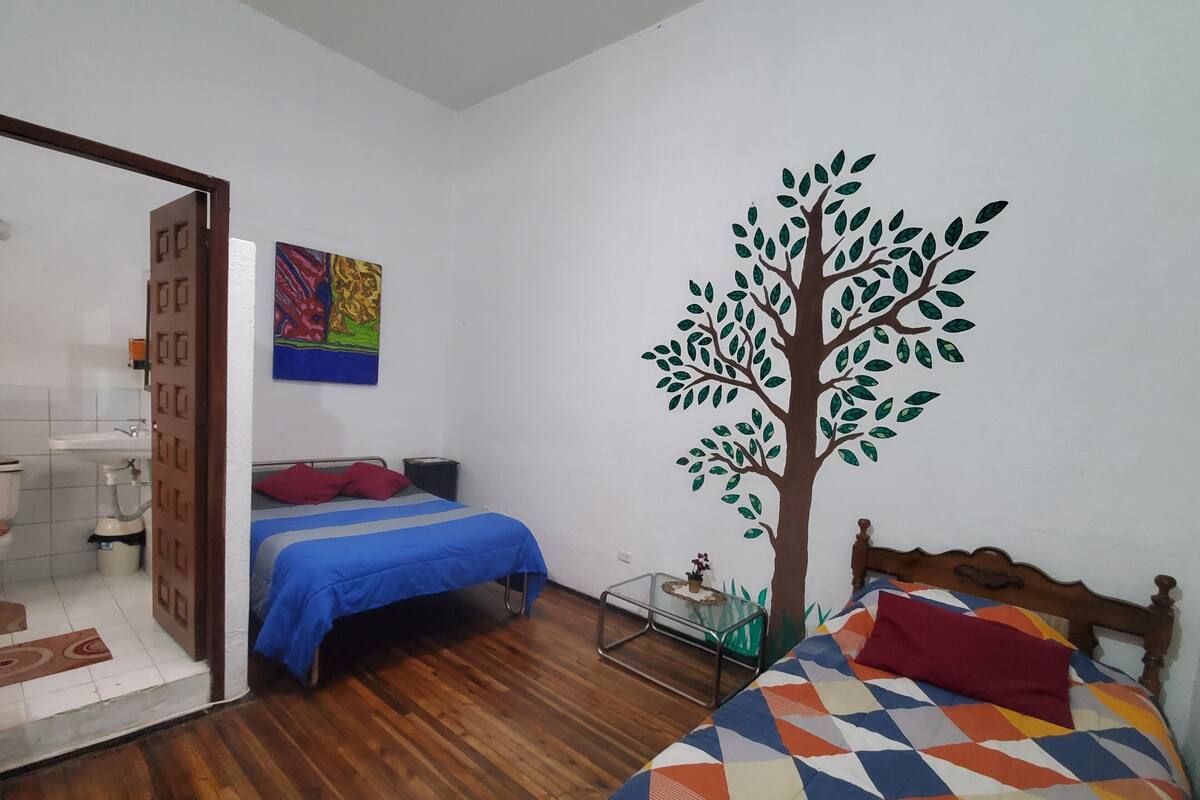 A bedroom with a wooden floor, a bed with a colorful geometric-patterned blanket, a wall mural of a tree, and a bathroom visible through an open door.