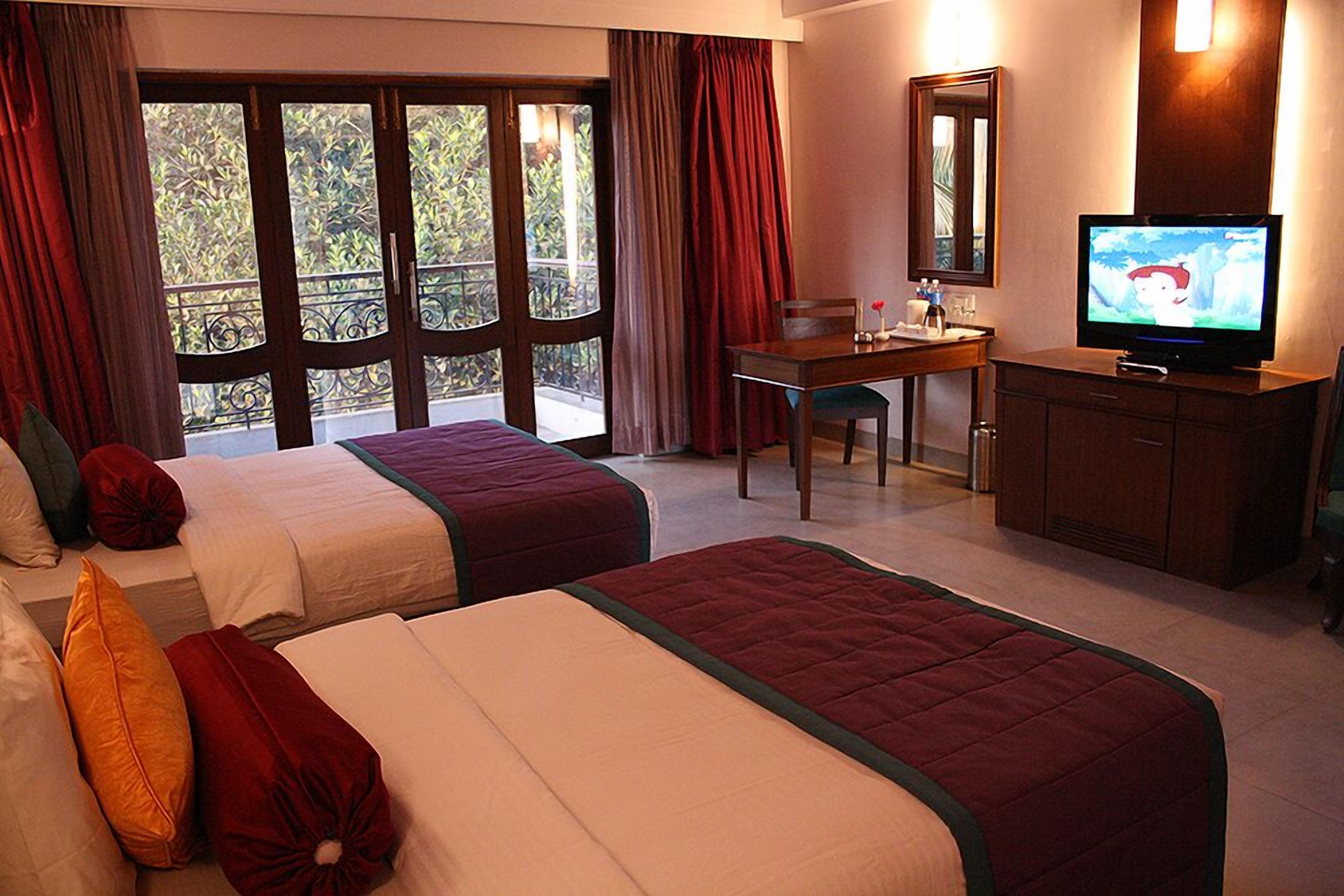 A hotel room with two beds, a desk, a television, and a balcony with curtains.