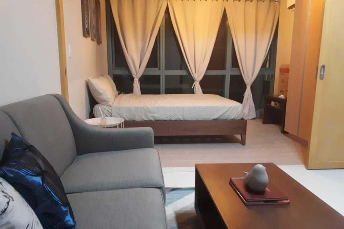 A room with a grey sofa, a small round table with a book and a decorative item, a bed with curtains, and a wooden door.