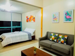 A bedroom with a bed, a gray sofa, and a small table with a plant. There are colorful paintings on the wall.
