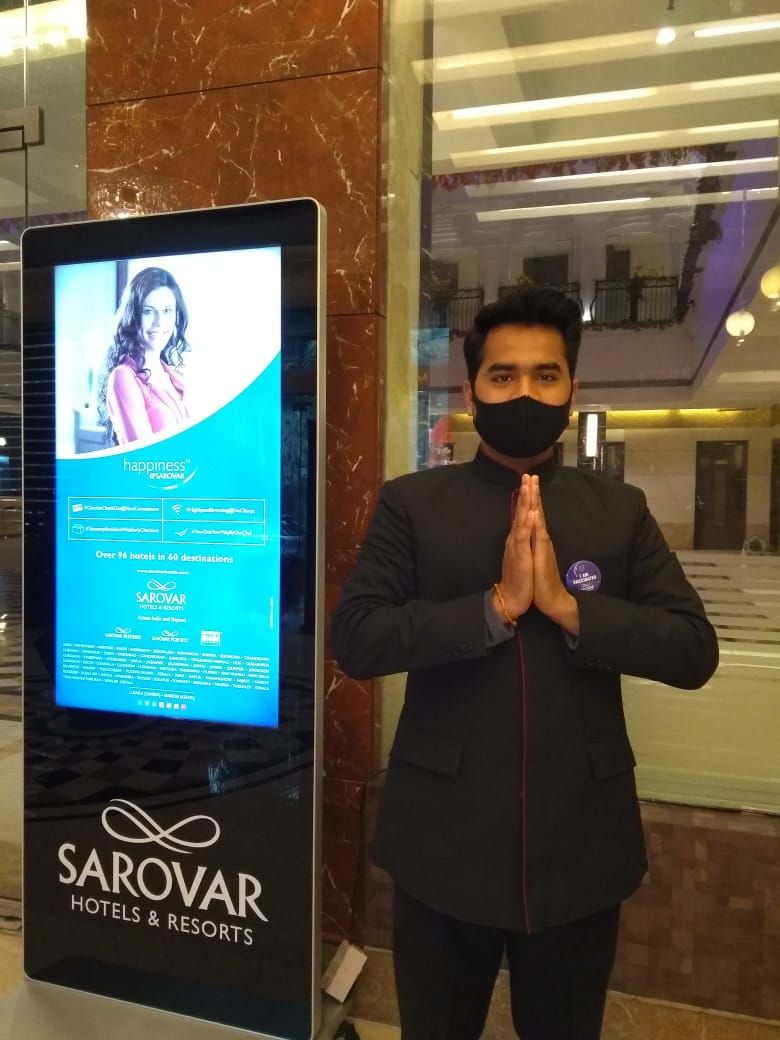 Ein Mann in einem schwarzen Anzug mit Abzeichen steht neben einer digitalen Anzeige, die Sarovar Hotels & Resorts wirbt.