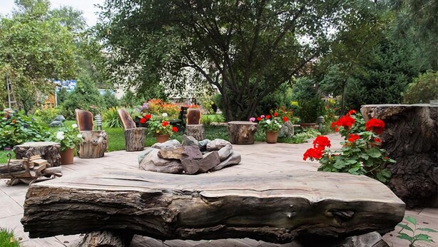 Uma área de estar ao ar livre rústica, com uma grande mesa de madeira cercada por elementos de pedra e madeira, situada num jardim exuberante com diversas plantas e flores.