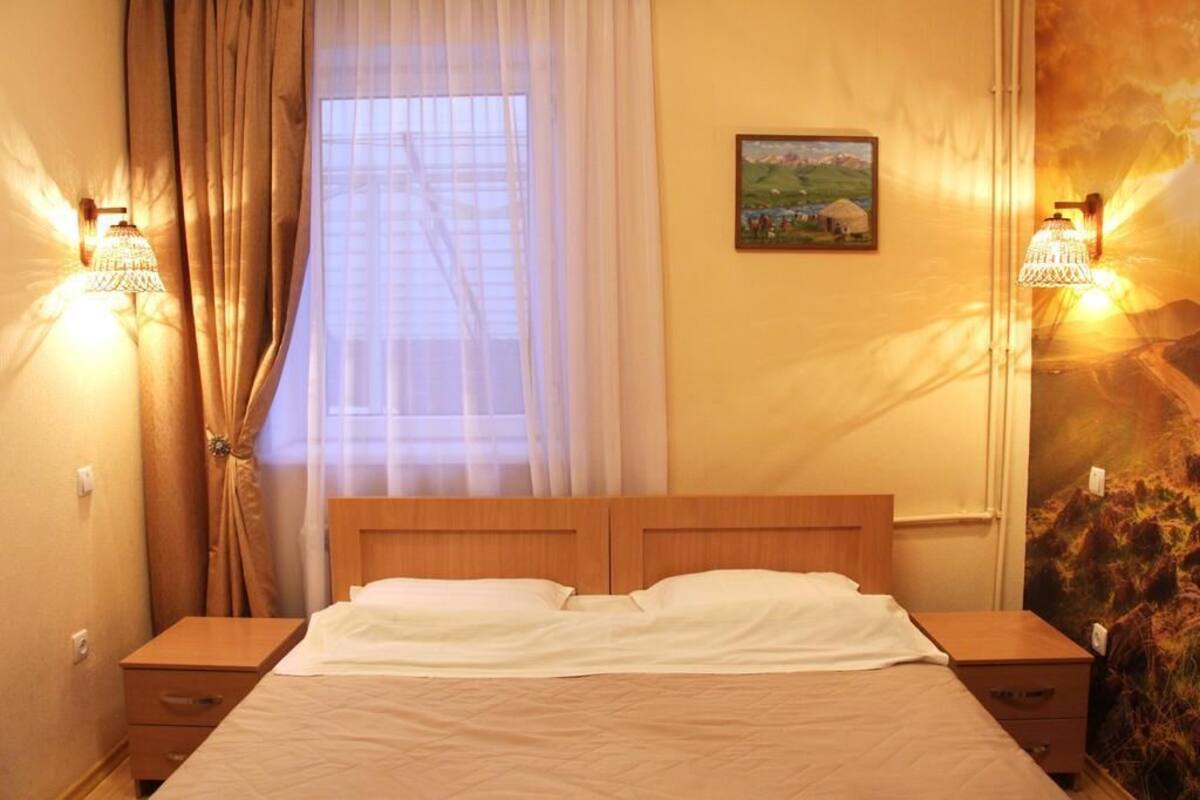 Una camera d'albergo con un letto in legno, due comodini, una finestra con tende leggere e lampade a parete.