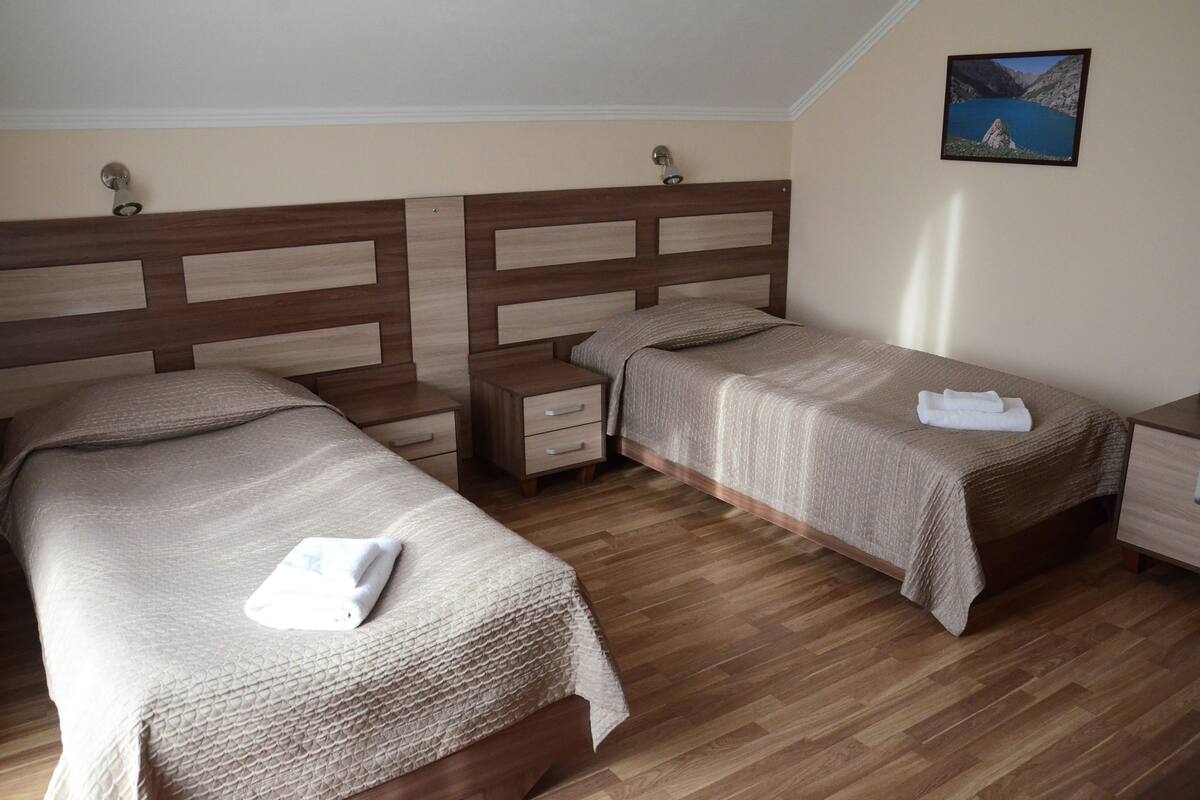 Camera d'albergo con due letti, testiere in legno, un comodino, un cassetto e un quadro appeso al muro.