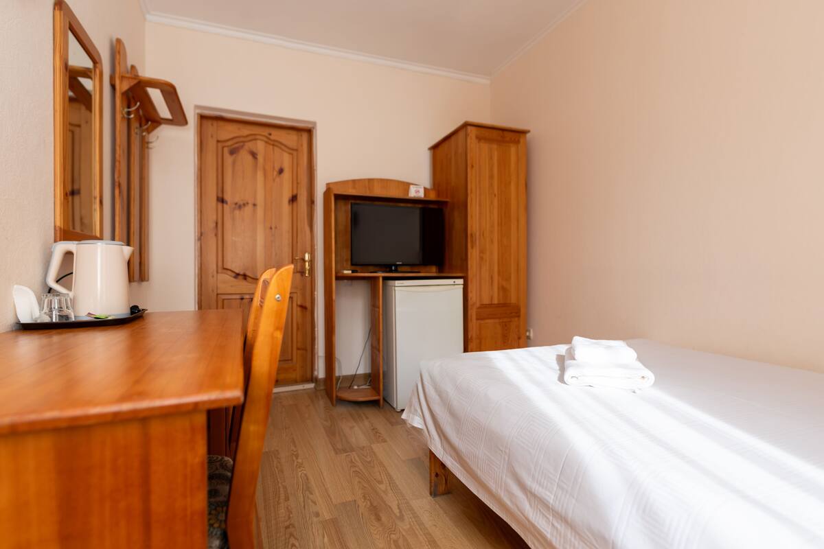 Una stanza con una scrivania in legno, una sedia, un letto con coperta bianca, una televisione e un piccolo frigorifero.