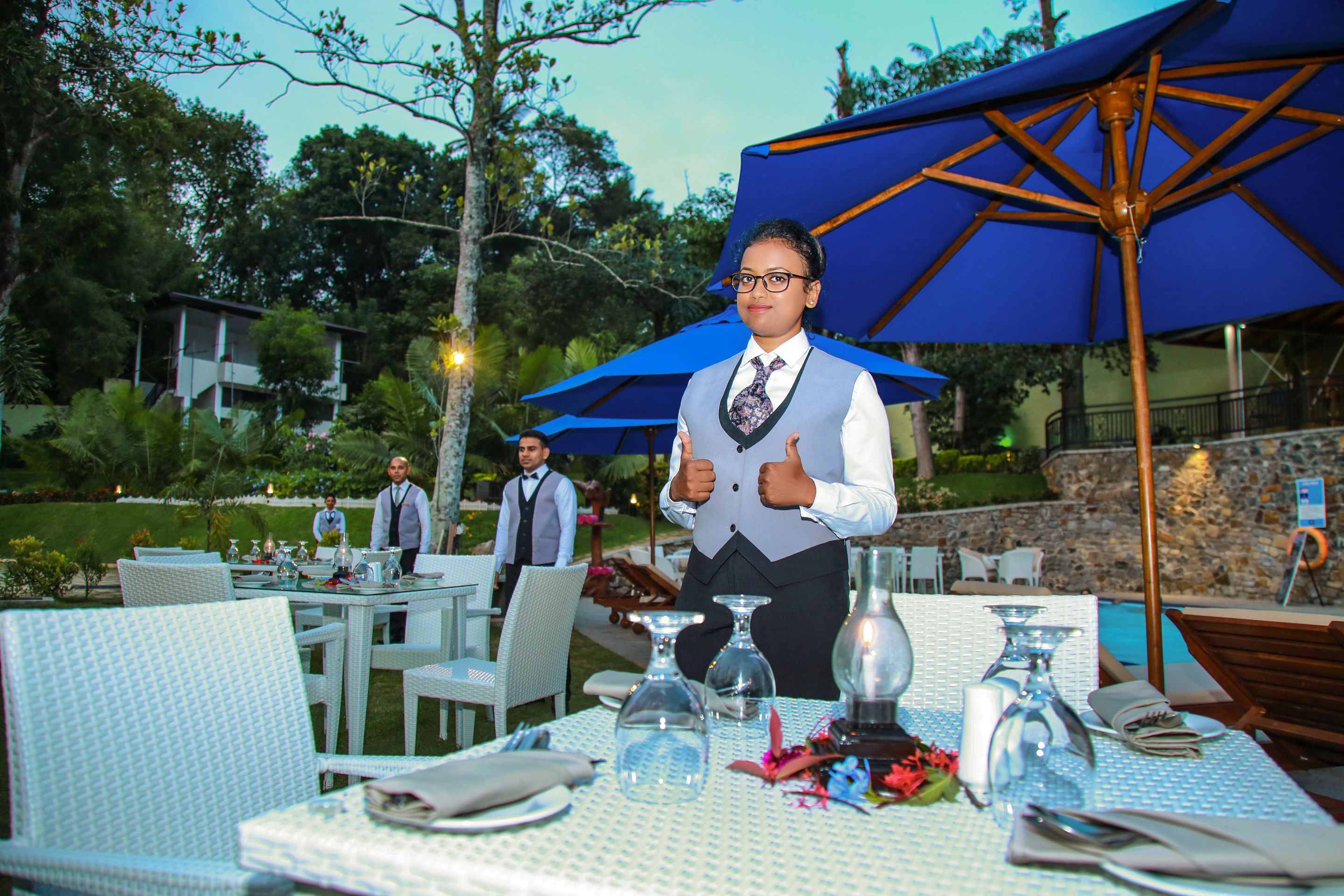 Un área de comedor al aire libre con sillas y mesas de mimbre blanco, sombrillas azules y una piscina al fondo.