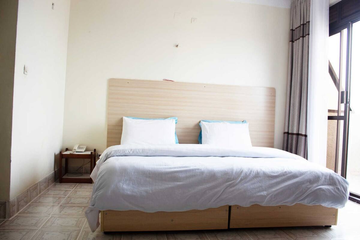 Une chambre d’hôtel avec un lit en bois, du linge de lit blanc, un téléphone sur la table de chevet et une fenêtre avec des rideaux.