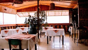Un restaurante con varias mesas cubiertas con manteles blancos y caminos de mesa estampados en rojo. Hay plantas en macetas y un gran jarrón decorativo en las mesas. El restaurante tiene un techo de madera con un diseño estampado y amplios ventanales que permiten la entrada de luz natural.