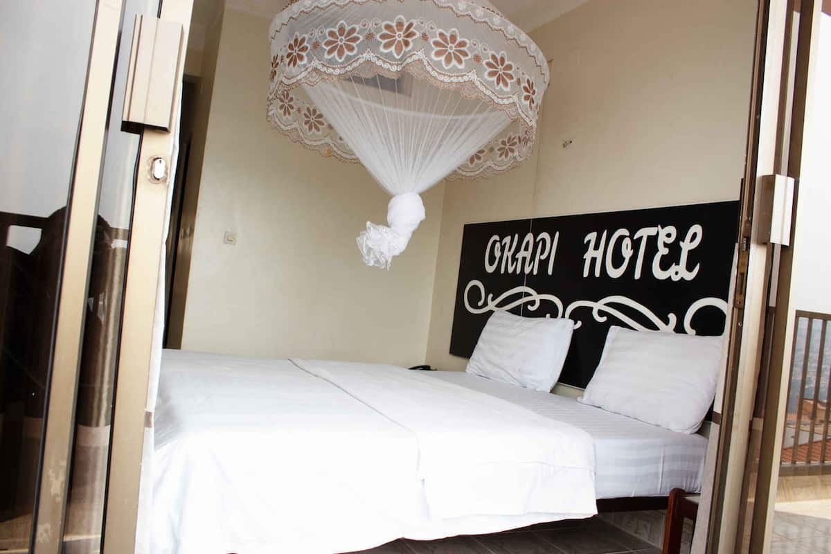 Un lit avec une literie blanche, un baldaquin orné de motifs floraux et une enseigne indiquant « OKAPI HOTEL ».