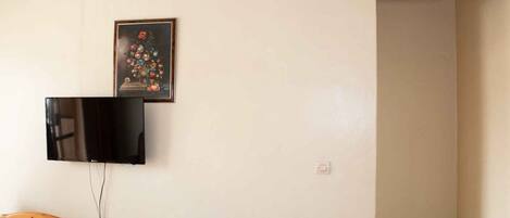 A room with a flat-screen TV mounted on the wall, a wooden chair, a small table, and a framed floral painting.