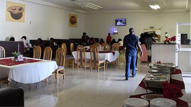 A dining area with tables and chairs, a television screen, and a counter with food items. - Ekamuti Town lodge (Ondangwa)