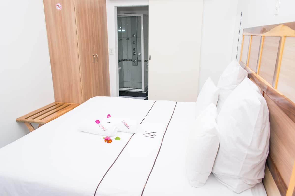 Een net opgemaakt bed met witte lakens en een houten hoofdbord, een houten kledingkast en een douchedeur van glas.