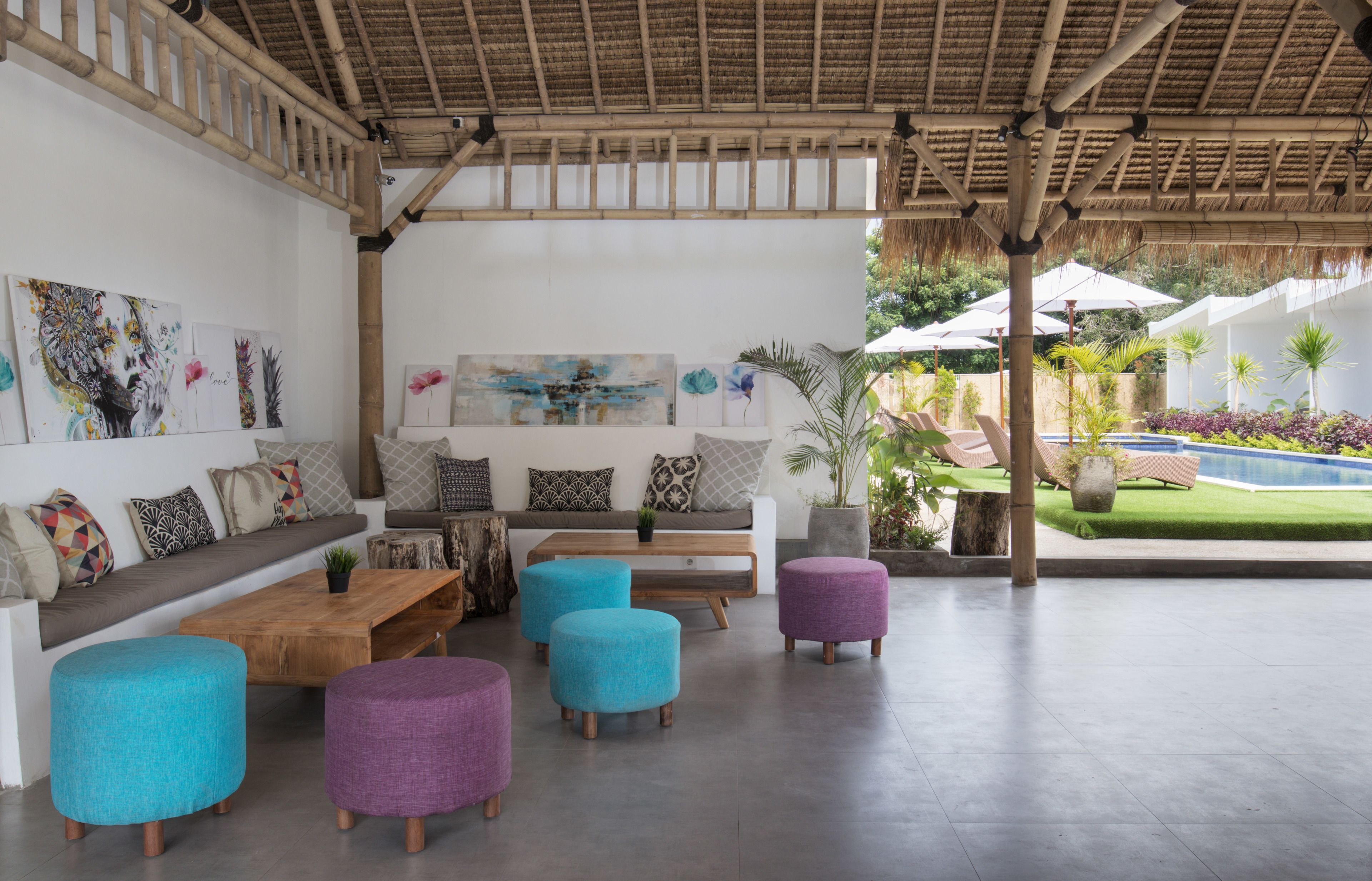 A covered outdoor seating area with a wooden bench, colorful ottomans, and artwork on the walls.