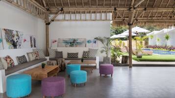 A covered outdoor seating area with a wooden bench, colorful ottomans, and artwork on the walls.