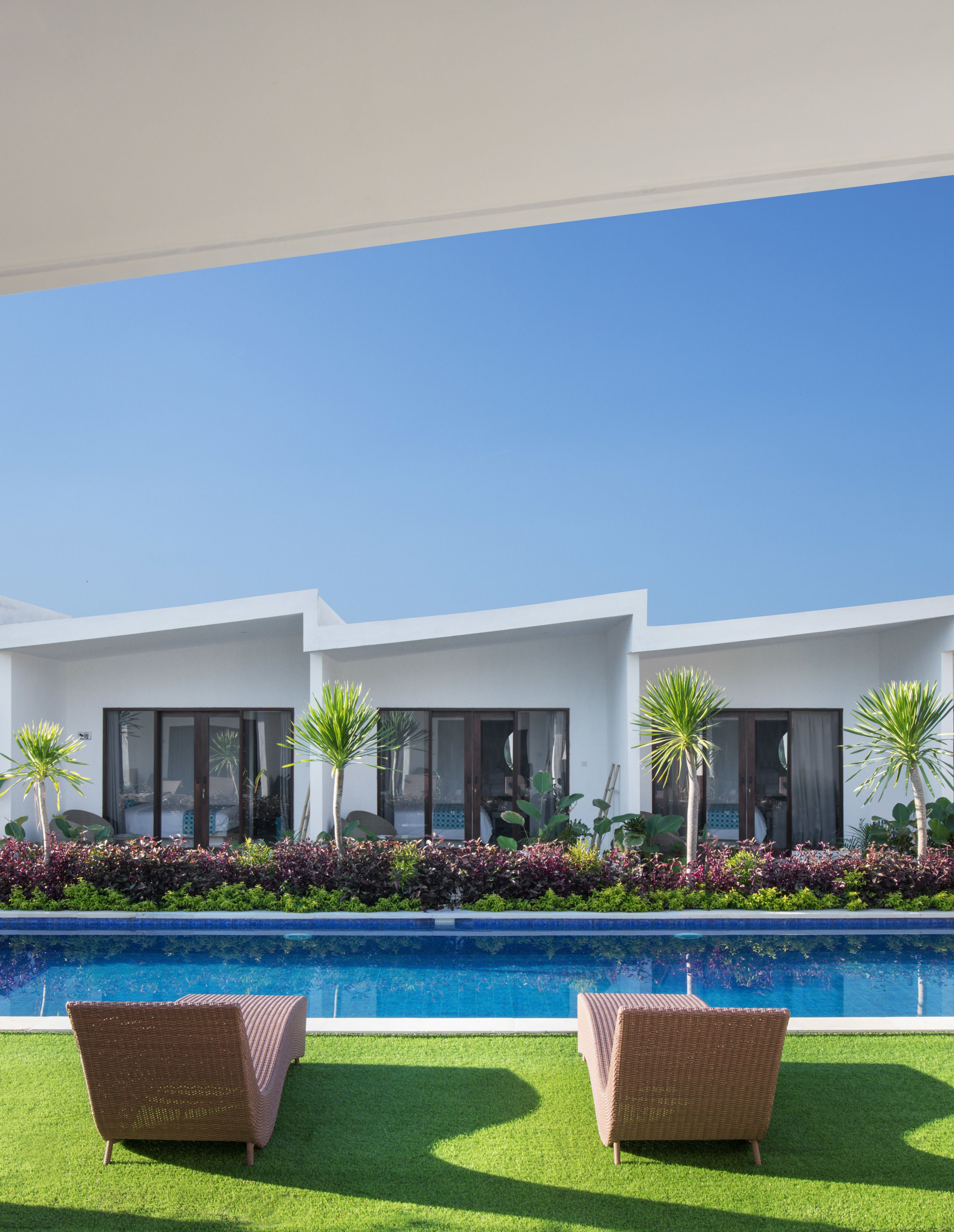 A modern building with a pool, two wicker chairs, and tropical plants.