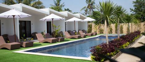 A pool area with lounge chairs and umbrellas, surrounded by palm trees and a clear sky.