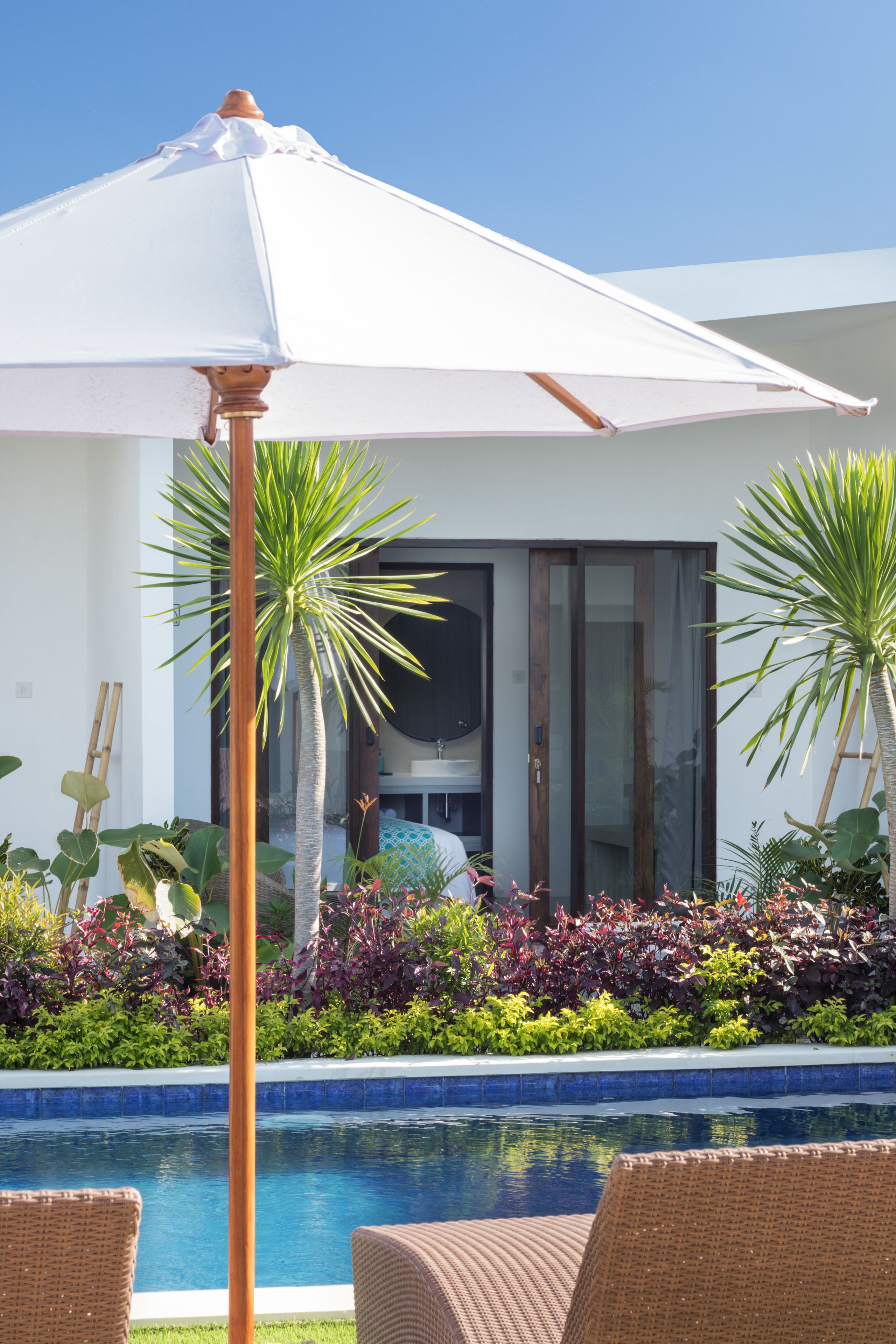 A pool area with a white umbrella, wicker lounge chairs, and tropical plants.