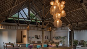 Un salón de planta abierta con mesas y bancos de madera, techo alto con vigas de bambú y lámparas esféricas colgantes.